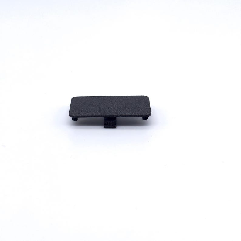 May include: A black plastic cover for a small electronic device. The cover has a textured surface and a small tab on the side.