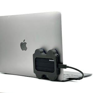 Könnte beinhalten: Ein silberner Laptop mit einer schwarzen externen Festplatte, die daran angeschlossen ist. Die Festplatte ist mit "SAMSUNG" beschriftet. Ein schwarzes Kabel verbindet die Festplatte mit dem Laptop.