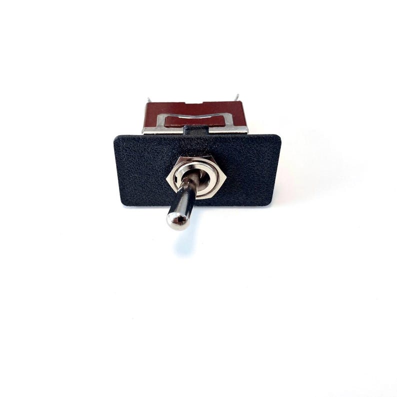 May include: A black toggle switch with a silver lever and a black plastic housing. The switch is mounted on a black rectangular plate.