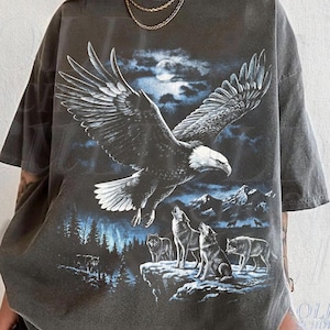 May include: A charcoal gray short-sleeve t-shirt featuring a graphic print of a bald eagle in flight above a mountain landscape with three wolves standing on a rocky cliff under a full moon. The design includes blue and white highlights.