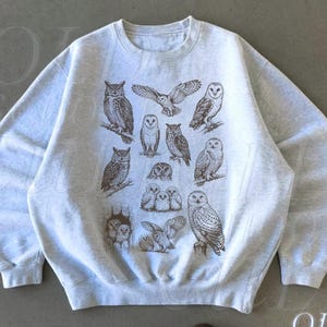 May include: Ash-colored sweatshirt featuring a brown illustration of various owl species. The design includes owls perched on branches and in flight. The text "OLD SCHOOL CULT" is visible in the lower right corner.