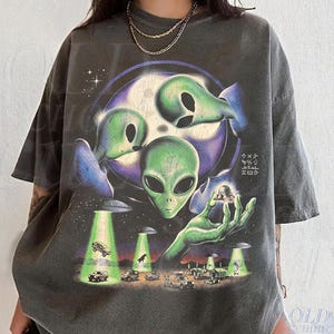 May include: A charcoal gray short-sleeve t-shirt featuring a graphic print of three green alien heads against a purple moon. A large green hand holds a small flying saucer above a landscape with multiple UFOs beaming light onto various vehicles.