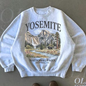 Op de afbeelding: Asgrijze crewneck sweatshirt met een Yosemite National Park-afbeelding. Het ontwerp toont een bergketen, waterval, rivier, beer en herten. De tekst "YOSEMITE" staat boven de afbeelding, met daaronder "NATIONAL PARK CALIFORNIA, USA".