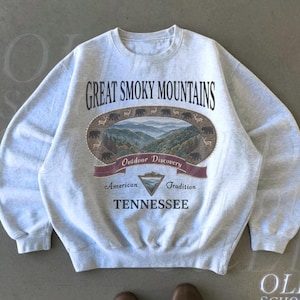 K&ouml;nnte beinhalten: Hellgraues Sweatshirt mit dem Schriftzug "Great Smoky Mountains" und einer Grafik einer Berglandschaft. Das Sweatshirt tr&auml;gt auch die Worte "Outdoor Discovery", "American Tradition" und "Tennessee".
