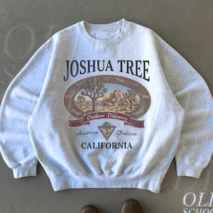 Pode incluir: Camisola cinza-claro com a inscrição "JOSHUA TREE" acima de um gráfico oval de uma paisagem desértica. O gráfico inclui as palavras "Outdoor Discovery", "American Tradition" e "CALIFORNIA". A camisola faz parte da coleção "OLD SCHOOL CULT".