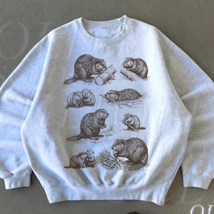 May include: Light grey sweatshirt with a detailed graphic of beavers in different poses. The design includes multiple beavers, some with logs and branches. The text "OLD SCHOOL CULT" is visible in the lower right corner.