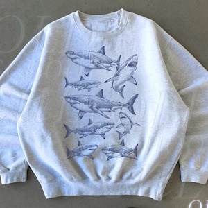 Puede incluir: Sudadera gris ceniza con un gráfico azul de varios tiburones. La sudadera tiene cuello redondo y mangas largas. El texto "OLD SCHOOL CULT" está impreso en blanco en la esquina inferior derecha. La palabra "Ash" está impresa en la esquina inferior izquierda.