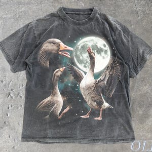 Pode incluir: Uma t-shirt de manga curta em cinzento-carvão com uma impressão gráfica de três gansos contra um céu noturno com uma lua cheia grande e estrelas. Os gansos estão representados em várias poses, com um deles a abrir as asas. O tecido tem um aspeto desbotado.