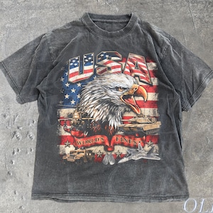 May include: Charcoal grey t-shirt with a graphic of a bald eagle and the American flag. The word "USA" is across the top, with "WORLD'S FINEST" below. The shirt has a vintage, distressed appearance. The text "OLD SCHOOL CULT" is at the bottom.