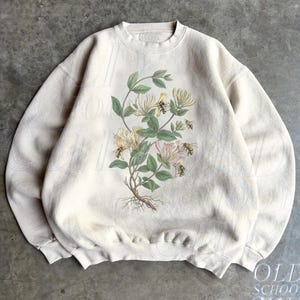 May include: A sand-colored crewneck sweatshirt featuring a botanical illustration of honeysuckle flowers with green leaves and several small bees. The design is centered on the chest, showcasing detailed floral stems and roots in soft tones.