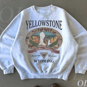 Może przedstawiać: Popielata bluza z nadrukiem w stylu vintage z Parku Narodowego Yellowstone. Projekt przedstawia owalną ilustrację z gejzerem, bizonami i tekstem "Yellowstone", "Outdoor Discovery" i "Wyoming".