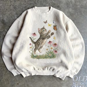 May include: A sand-coloured crewneck sweatshirt featuring a graphic print of a tabby kitten reaching for butterflies amidst pink and white wildflowers. The kitten is depicted in a playful pose, surrounded by several small, colourful butterflies.