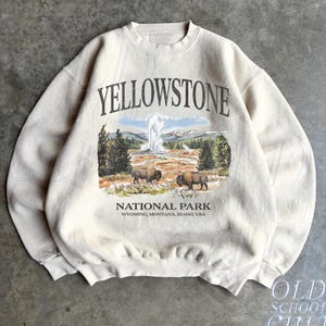 May include: A sand-colored crewneck sweatshirt featuring a graphic print of Yellowstone National Park. The design includes the text YELLOWSTONE NATIONAL PARK WYOMING, MONTANA, IDAHO, USA, a geyser, pine trees, and two brown bison in a landscape.