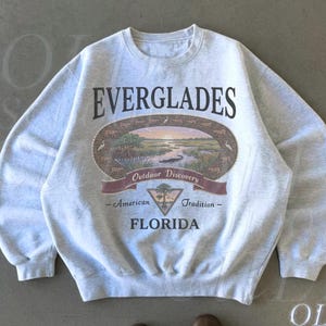 May include: Light grey crewneck sweatshirt with the word "EVERGLADES" in black block letters above a landscape graphic. The graphic includes a river scene with a sunset, and the words "Outdoor Discovery", "American Tradition", and "FLORIDA".