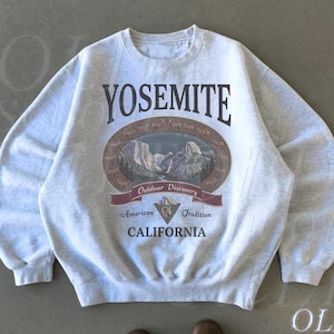Puede incluir: Sudadera gris ceniza con un diseño del Parque Nacional de Yosemite. El diseño presenta la palabra "YOSEMITE" sobre una ilustración ovalada de un paisaje montañoso, con el texto "Outdoor Discovery, American Tradition, CALIFORNIA."