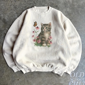 Puede incluir: Una sudadera de cuello redondo color arena con un estampado gráfico de un gatito atigrado marrón sentado entre flores de equinácea rosas con una mariposa monarca sobrevolando. El jersey de manga larga tiene cuello, puños y dobladillo acanalados.