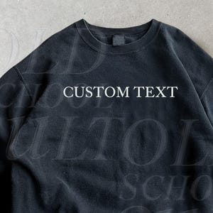 Może przedstawiać: Czarna bluza z okrągłym dekoltem z białym napisem "CUSTOM TEXT". Ubranie ma wyblakły wygląd. Słowo "Black" jest widoczne w lewym dolnym rogu. Codzienny, swobodny element garderoby.