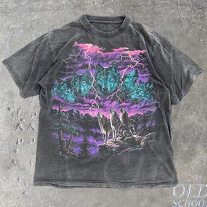 May include: A charcoal grey short-sleeve t-shirt featuring a graphic print of multiple wolves against a backdrop of purple clouds and lightning. The design includes three wolves standing on a rocky mountain ledge below the wolf heads.