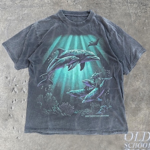 Op de afbeelding: Een antracietgrijs T-shirt met korte mouwen en een grafische print van dolfijnen die in turquoise water boven koraalriffen zwemmen. De tekst DEEP WATER EXPLORATIONS staat in kleine witte letters rechtsonder in het grafische ontwerp.
