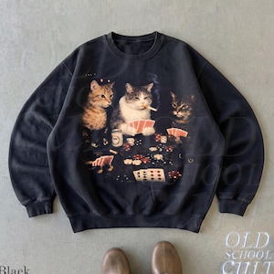 Könnte beinhalten: Ein schwarzes Sweatshirt mit Rundhalsausschnitt und einem Grafikdruck von drei Katzen, die an einem Tisch sitzen und Karten spielen. Die mittlere Katze hat eine Zigarette im Mund und der Tisch ist mit Spielkarten und Pokerchips bedeckt.