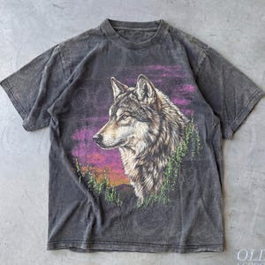 May include: Dark gray t-shirt featuring a detailed illustration of a wolf's head against a purple and orange sunset backdrop. The wolf is depicted in shades of brown and gray, with green trees in the foreground. The shirt has a vintage, worn look.