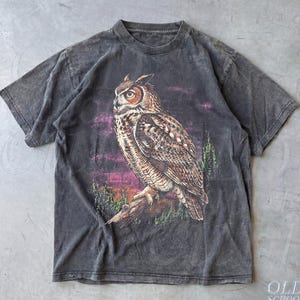 May include: Dark gray t-shirt featuring a detailed illustration of a Great Horned Owl perched on a branch. The owl is depicted with brown and white feathers against a backdrop of a sunset. The shirt has a vintage, distressed look.