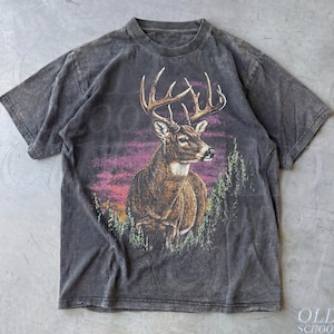 Peut inclure: Un t-shirt gris anthracite avec un cerf majestueux orné de grands bois, sur fond de coucher de soleil aux teintes violettes et orangées. Des arbres verts encadrent le motif. Le t-shirt a un aspect vintage et délavé.
