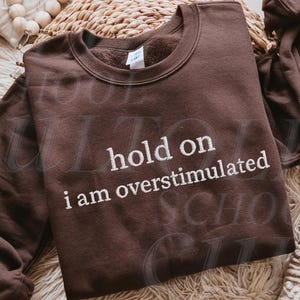 Puede incluir: Sudadera de color chocolate oscuro con la frase bordada "hold on i am overstimulated" en hilo blanco. La sudadera se muestra sobre una superficie tejida, mostrando su estilo casual y diseño cómodo. El color del hilo es blanco.