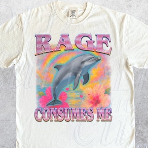 May include: Ivory-colored t-shirt with a graphic design featuring a dolphin leaping from the water, surrounded by a rainbow and flowers. The text "RAGE CONSUMES ME" is printed above and below the dolphin in a pink and purple font.