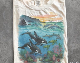 May include: An ivory t-shirt featuring a graphic print of an underwater scene with two orcas, sea turtles, and coral reefs, topped by a coastal view with dolphins swimming in the ocean under a sunset sky. The shirt has a Comfort Colors brand label.
