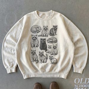 May include: Sand-colored sweatshirt featuring a repeating pattern of raccoon illustrations in various poses. The graphic is in black and white. The sweatshirt has a crew neck and long sleeves. The text "OLD SCHOOL CULT" is printed on the lower right.