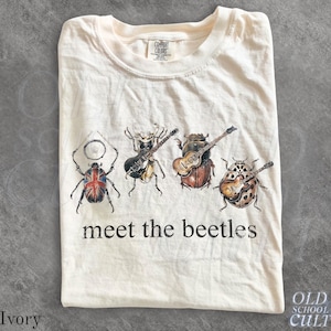 Op de afbeelding: Een ivoorkleurig t-shirt met een afbeelding van vier kevers die muziekinstrumenten bespelen, waaronder een drumstel, een zwarte elektrische gitaar, een akoestische gitaar en een bruine elektrische gitaar. De tekst "meet the beetles" staat onder de insecten.