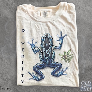 May include: An ivory t-shirt featuring a graphic of a blue and black poison dart frog with the word DIVERSITY printed vertically in black capital letters on the left side. The shirt has a crew neckline and a Comfort Colors brand label at the neck.