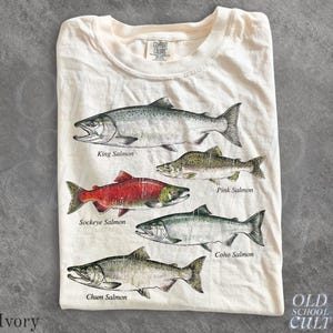 以下が含まれることがあります： アイボリー色のTシャツで、キングサーモン、ピンクサーモン、ベニザケ、ギンザケ、シロザケという5種類のサケのグラフィックプリントが施されています。Tシャツはクルーネックで、首元にComfort Colorsのブランドラベルが付いています。