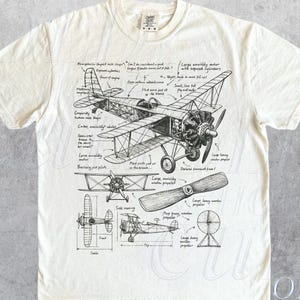 May include: Ivory t-shirt with a detailed black and white diagram of a vintage biplane, including labeled parts and a propeller. The shirt has a classic crew neck and short sleeves. The text "OLD SCHOOL CULT" is printed on the bottom right.