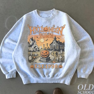 May include: A light grey crewneck sweatshirt featuring a graphic print with the text HALLOWEEN TOWN EST. 1998. The illustration depicts a spooky scene with a large jack-o'-lantern, a church, tombstones, bats, and a full moon in orange and black.