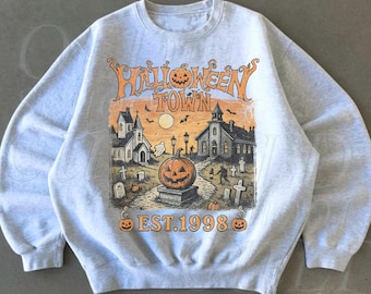 May include: A light gray crewneck sweatshirt featuring a graphic print with the text HALLOWEEN TOWN EST. 1998. The illustration depicts a spooky scene with a large jack-o'-lantern, a church, tombstones, bats, and a full moon in orange and black.