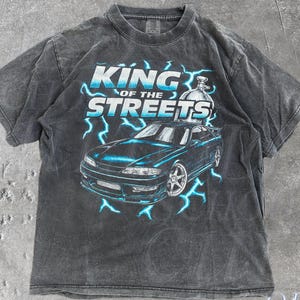 May include: A charcoal gray t-shirt featuring a graphic of a sports car with blue lightning bolts and the text "KING OF THE STREETS" in blue and white. The shirt has a vintage, washed look and the words "OLD SCHOOL CULT" are printed on the bottom right.
