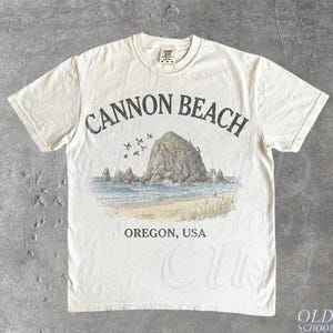 Könnte beinhalten: Ein elfenbeinfarbenes Kurzarm-T-Shirt mit einem Grafikdruck von Haystack Rock am Cannon Beach, Oregon. Der Text CANNON BEACH steht in gebogener Schrift über der Illustration und OREGON, USA in gerader Schrift unter der Küstenszene.