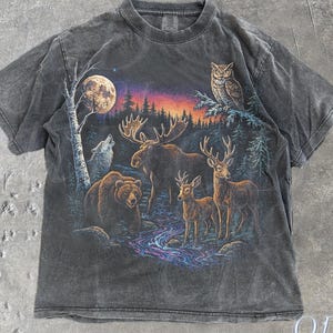 Puede incluir: Camiseta gris oscuro con una escena de vida silvestre que presenta un oso, un alce, ciervos, un lobo y un búho bajo la luna llena. El gráfico incluye una colorida puesta de sol y un bosque. El texto "OLD SCHOOL CULT" está en la parte inferior derecha.