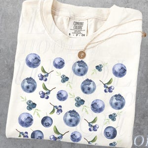Op de afbeelding: Een wit T-shirt met een blauw bosbessenpatroon en de tekst "OLD SCHOOL CULT" gedrukt in de rechterbenedenhoek.