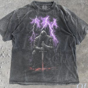 Könnte beinhalten: Pfefferfarbenes T-Shirt mit einer Grafik eines knienden Ritters mit einem Schwert unter violetten Blitzen. Das Shirt hat einen Vintage-Wascheffekt. Der Text "OLD SCHOOL CULT" ist unten rechts aufgedruckt.