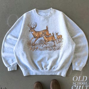 May include: An ash grey crewneck sweatshirt featuring a detailed graphic print of three deer in a natural landscape. The illustration depicts a large buck with antlers, a doe, and a fawn running through tall grass and scattered oak leaves.