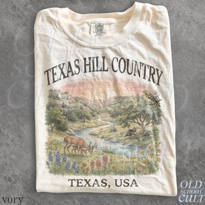 Könnte beinhalten: Ein elfenbeinfarbenes T-Shirt mit einem Grafikdruck einer Texas Hill Country Landschaft. Das Design zeigt einen gewundenen Fluss, einen Hirsch, einen Vogel und Wildblumen. Der Text lautet TEXAS HILL COUNTRY oben und TEXAS, USA unten.