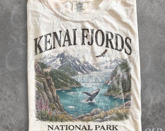 Könnte beinhalten: Ein elfenbeinfarbenes T-Shirt mit einer Grafik des Kenai Fjords Nationalparks in Alaska. Das Design zeigt Berge, einen Gletscher, einen Wal im Wasser, eine Bergziege auf einer Klippe und einen Adler mit dem Text Kenai Fjords National Park Alaska, USA.