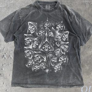 以下が含まれることがあります： 複数のフード付きの頭蓋骨が描かれた、濃いグレーのTシャツ。デザインはグレーと白の濃淡で、印象的なコントラストを生み出しています。「OLD SCHOOL CULT」の文字が右下に表示されています。