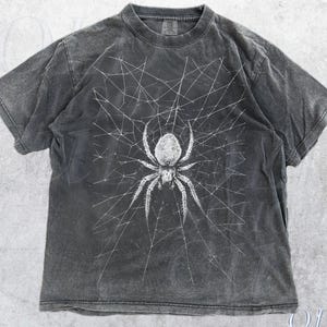 May include: A dark grey t-shirt with a white spider and web design. The tee has a vintage, worn appearance. The text "OLD SCHOOL CULT" is printed on the bottom right corner.