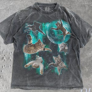 May include: Dark gray t-shirt featuring a graphic of cats floating in space with a teal and white aurora borealis effect. The shirt has a vintage, washed look. The text "OLD SCHOOL CULT" is printed on the bottom right.