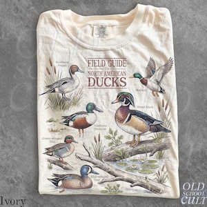 Peut inclure: Un t-shirt ivoire avec une impression graphique intitulée Field Guide to North American Ducks. L'illustration montre un pilet, un colvert, un canard branchu, un canard souchet, une sarcelle à ailes vertes et une sarcelle à ailes bleues.