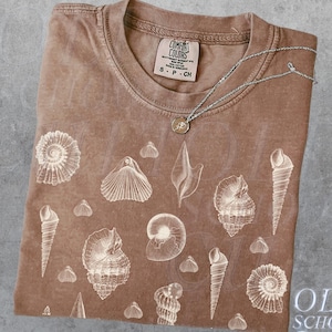 以下が含まれることがあります： 様々な貝殻の白いプリントが施された茶色のTシャツ。シャツには「Comfort Colors」と「S - P - CH」と書かれたタグが付いています。